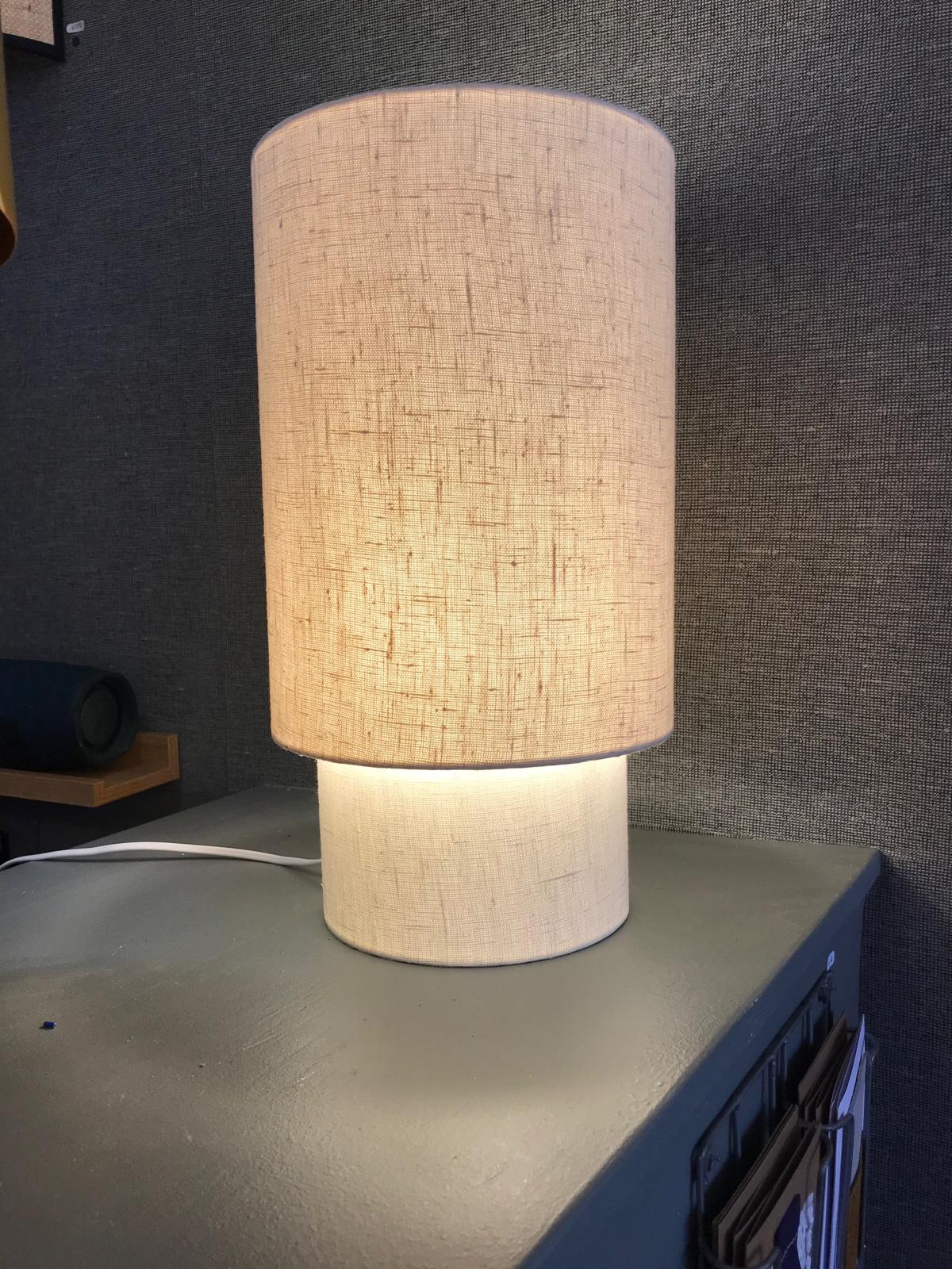 Lampe cylindrique claire allumÃ©e posÃ©e sur un meuble devant un mur textile sombre